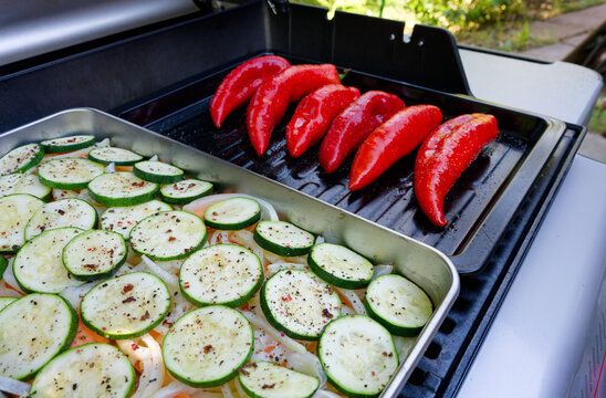 Fresh red pointed peppers and seasoned zucchini slices are cooking on a barbecue grill. A close-up of a gas grill loaded with fresh vegetables.