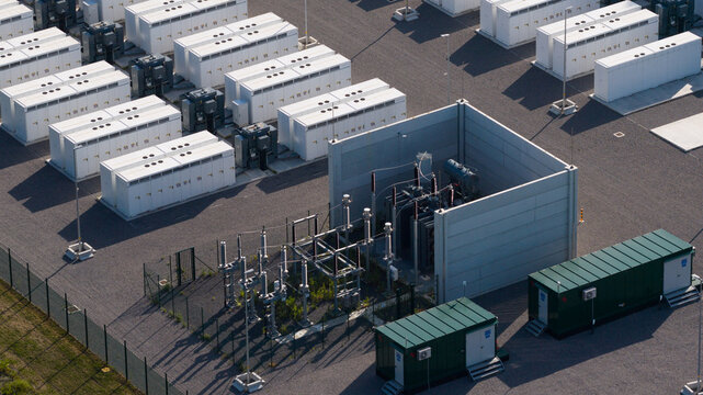 Aerial view of a large-scale battery energy storage system with rows of white container units and electrical transformers on a gravel site in Le Roeulx, Wallonia, Belgium.