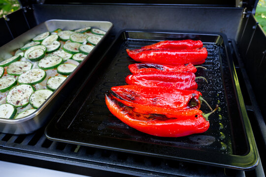 Fresh red pointed peppers and seasoned zucchini slices are cooking on a barbecue grill. A close-up of a gas grill loaded with fresh vegetables.