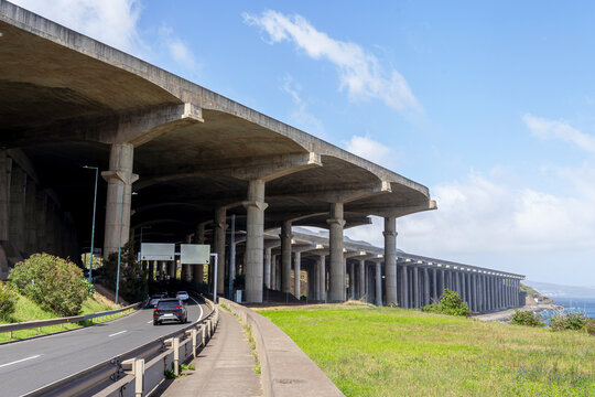 A view of the massive concrete pillars supporting the Madeira Airport runway extension in Santa Cruz, Portugal, with cars driving on the road beneath the structure.
