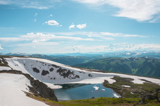 Scenic sunlit landscape with alpine lake in rocky snowy cirque near stone hill top during thaw. Ice floats in mountain lake among rocks and snows with view to forest mountain range under cloudy sky.