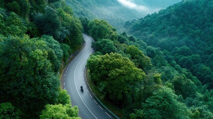 Fototapeta premium Motorcycle riding on winding mountain road through dense green forest with distant mist aerial view