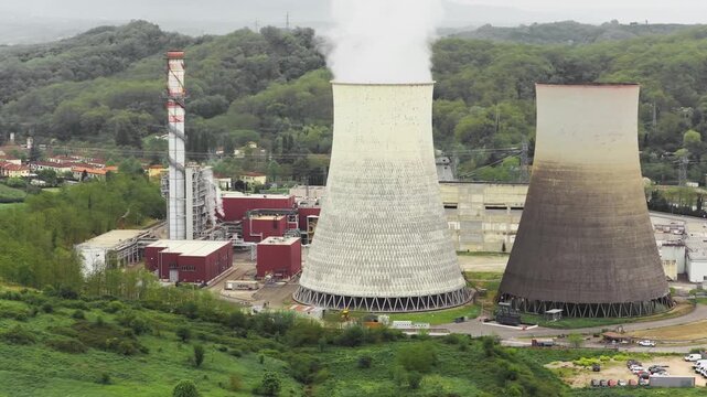 Aerial view of the Centrale Termoelettrica Santa Barbara Enel with large cooling towers emitting steam against a backdrop of lush green hills in Santa Barbara, Toscana, Italy.