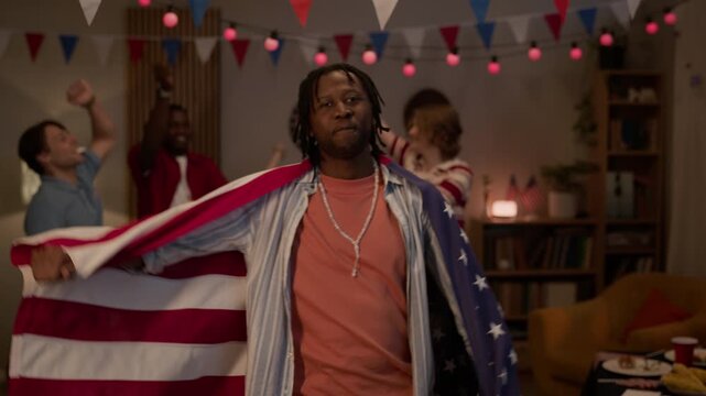 Medium portrait of young Black man wrapped in USA flag dancing with multiethnic friends at home party. Footage is ideal for Independence Day marketing campaigns and patriotic holiday promotions