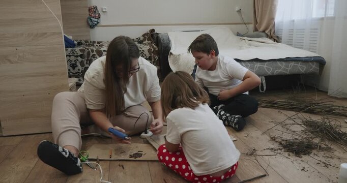 Caring mother with her son and daughter sitting on the floor, creating a handmade project together from natural materials and using a hot glue gun for a school assignment or creative hobby