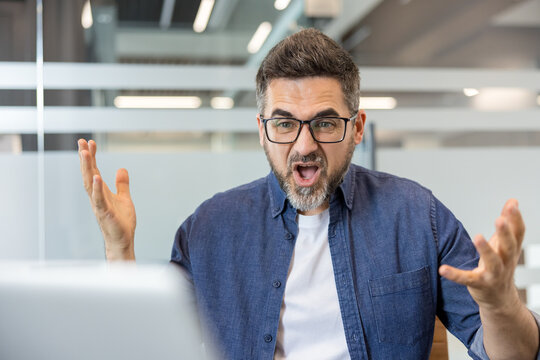 Angry businessman expressing strong emotions and stress, shouting and gesturing with arms outstretched in an office setting, feeling immense frustration and annoyance while working