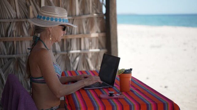 Woman engaged in video chat on computer and using phone on a beach during her vacation in a tropical location.