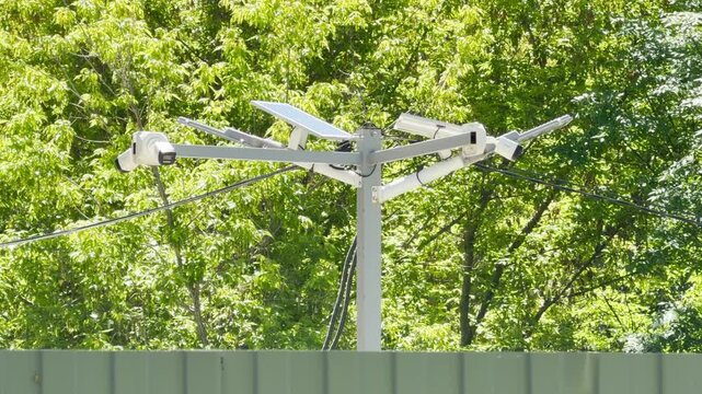 A central pole supports multiple security cameras and small solar panels. The apparatus stands as a monitoring device against a backdrop of dense, sunlit green trees. It blends technology with nature.