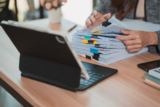 A close-up image of a businesswoman's hands rummaging through a large stack of documents on her desk, organizing papers and files in her office using a digital tablet.