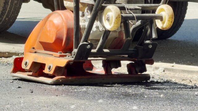A bright orange mechanical plate compactor rests on a patch of freshly laid asphalt. It is used to flatten and firm the surface. The machine stands on a busy urban road.