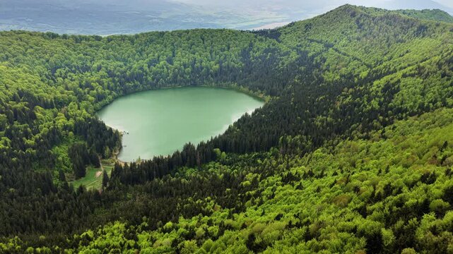 Aerial drone view of a round crater lake from a forested slope with distant ridges, Lacul Sfanta Ana, Harghita, Romania.