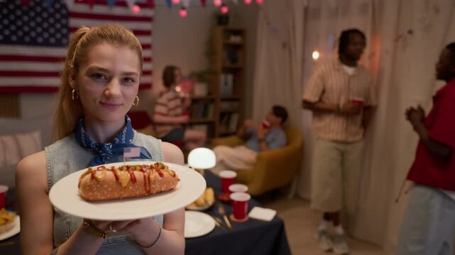 Chest up portrait of Caucasian young adult woman putting USA flag toothpick into hot dog at party with multiethnic friends in background. Ideal for 4th of July marketing and holiday food promotions