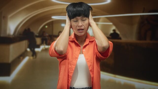Woman covering ears with hands, grimacing in an orange shirt and short bangs, standing in a modern metro station building concourse with ring lights and counters; stress.