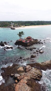 Aerial view of Seenigama Muhudu Dewalaya temple on a rocky island surrounded by turquoise ocean waves and a sandy coastline in Peraliya, Sri Lanka.