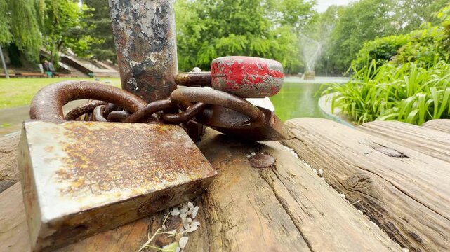 A rusty wedding padlock and a red heart lock are chained to a post. They sit on a wooden pier overlooking a green park pond with a fountain.