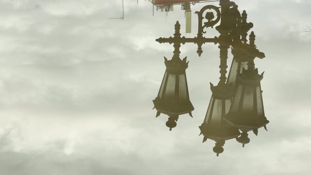 An ornate vintage street lamp reflects clearly in a rainwater puddle on the ground. The overcast cloudy sky fills the background of the water surface. It creates a calm and moody atmosphere.