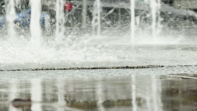Water jets spray forcefully from a ground fountain. The heavy splashes cover a wet concrete plaza. A blurry white car passes by on the street in the background.