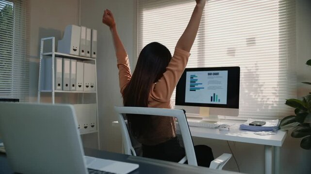 Rear view of a successful asian business woman finishing her work, stretching her arms, and feeling happy while sitting at her desk