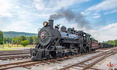 A steam locomotive moves through a train station on a sunny day. The engine releases smoke as it prepares for a journey. The mountains in the background add to the rural scene. © Greg Kelton