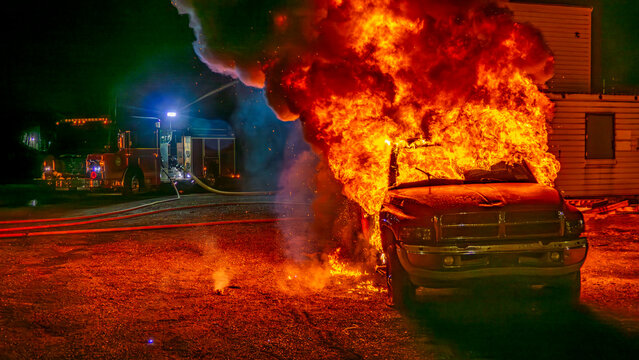 Flames rise high as a truck catches fire during a training drill for firefighters at night. The scene shows the truck fully engulfed in fire. Firefighters respond quickly to control the blaze.