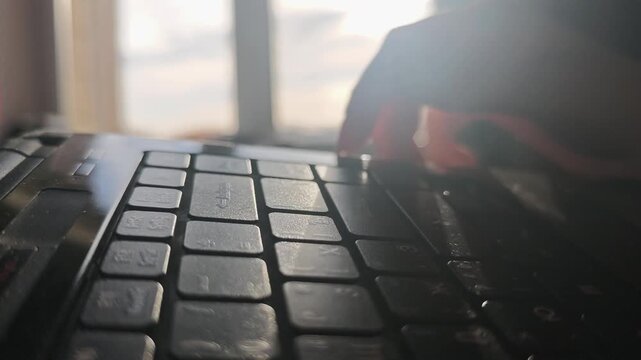 Close-up of hand pressing laptop keyboard keys while sunlight streams through window, creating a warm glow on the surface of the laptop in a cozy indoor setting