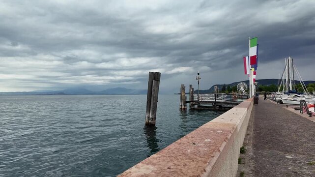 Bardolino, Italy - May 14, 2026 - Scenic Italian lakefront promenade with boats and flags under cloudy skies