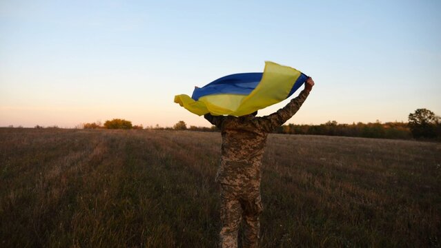 Soldier of ukrainian army running with raised flag Ukraine above his head at field. Young male military in uniform jogging with blue-yellow banner at meadow. Victory against russian aggression