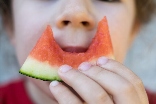 Front view close-up of a kid holding a triangular slice of watermelon with a visible crescent-shaped bite mark.