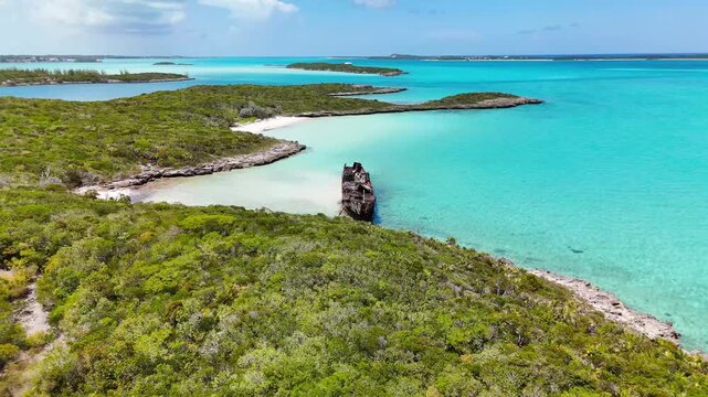 Aerial view of a rusty shipwreck on the white sand beach of Crab Cay surrounded by turquoise water and lush vegetation in George Town, Exuma, The Bahamas.