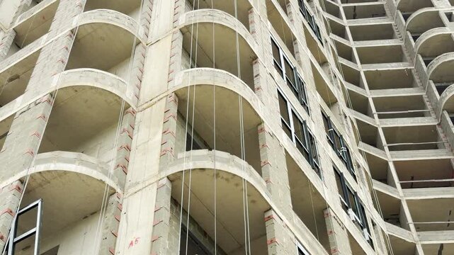 A raw concrete apartment building stands under development. The unfinished facade features curved balconies and new windows at a city construction site.