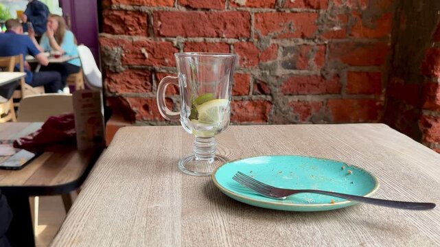 Empty turquoise plate with fork, glass tumbler holding lemon slice on linen tablecloth against exposed brick, intimate cafe vibe