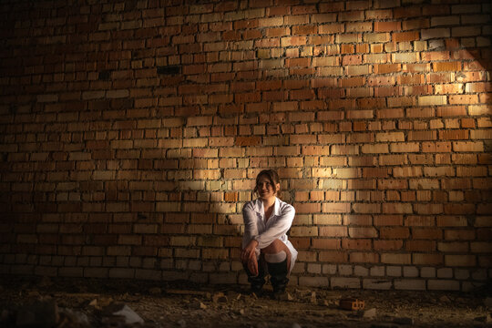 Young woman in white dress and high heels squats on uneven ground in an industrial setting with exposed brick walls and scattered debris in the background