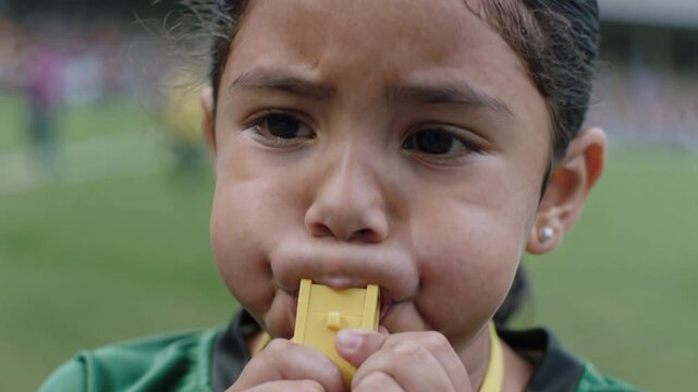 A young girl blows a yellow whistle with determination, her cheeks puffed out in concentration. She wears a green shirt with a yellow lanyard, suggesting a sporting event or role as a referee