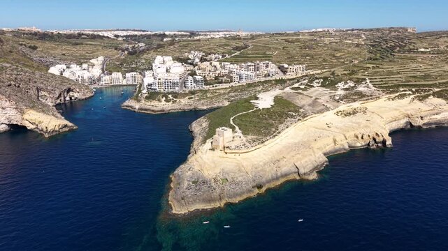 Aerial view of Xlendi Tower on the rocky cliffs overlooking the turquoise waters of Xlendi Bay with white buildings and terraced hills in Munxar, Gozo Region, Malta.