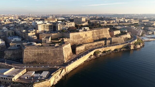 Aerial view of the massive limestone bastions and historic fortifications along the coastline of the old town during golden hour in Valletta, Port Region, Malta.