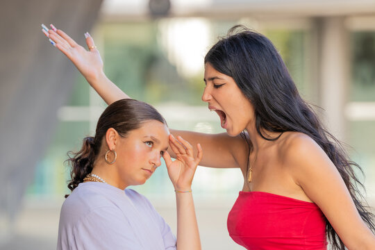 Young hispanic woman in red top yelling and gesturing at unbothered friend outdoors. Conflict, drama, and attitude concept