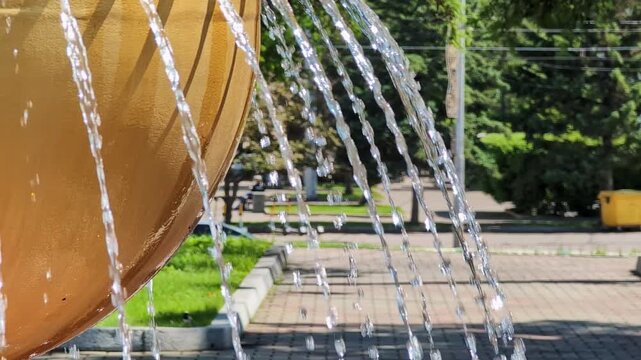 Golden spherical fountain featuring multiple streams of water arching outward in a park setting. Surrounded by paved walkways and green grass, the scene captures bright sunlight reflecting on the.