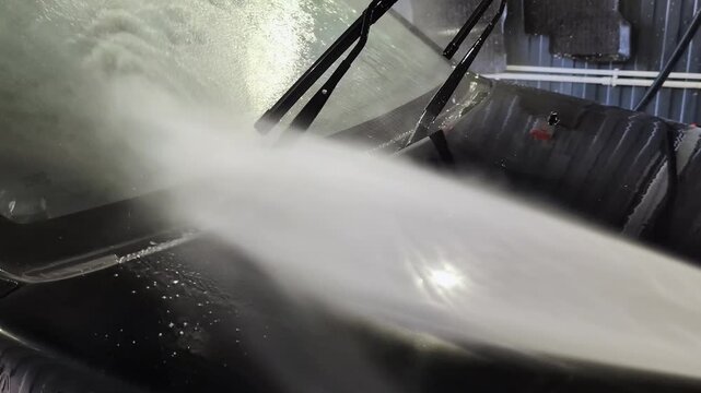 Black vehicle being cleaned with powerful jets of water spraying across the hood and windshield. Water droplets visible on the shiny surface and windshield wipers raised during the washing process.
