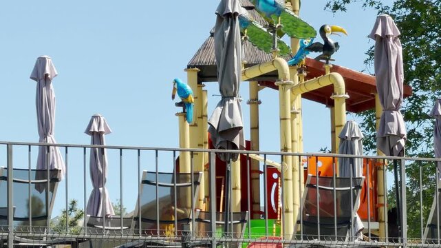 Colorful bird statues sit atop a bamboo-style play structure. Closed patio umbrellas and lounge chairs are arranged on the terrace. The setting is bright and sunny outdoors.
