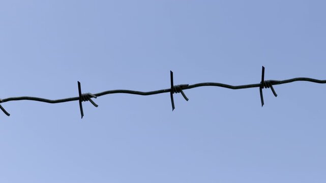 A single strand of barbed wire stretches horizontally across a clear blue sky. The sharp metal barbs contrast with the smooth, open background. The composition is simple and stark.