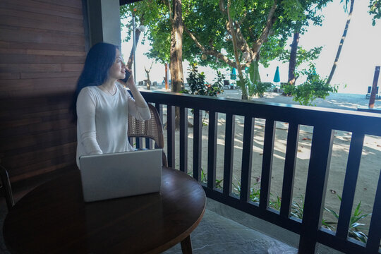 Young Asian woman talking on mobile phone while working on laptop from a tropical resort beach balcony. Digital nomad or remote worker concept.