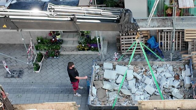 Construction worker operates crane to load debris into a dumpster for removal. A worker supervises a crane lifting rubble into a large container, signifying demolition or renovation progress.