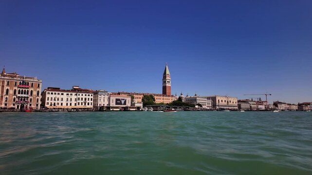 A wide view of the Venice skyline featuring St Mark Campanile and historic buildings. The city landscape is seen from the water of the Venetian lagoon on a clear sunny day.