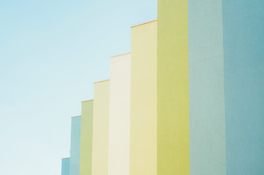 Low-angle view of a row of stepped building facades painted in soft pastel hues including blue, green, yellow and cream, set against a pale clear sky. Clean