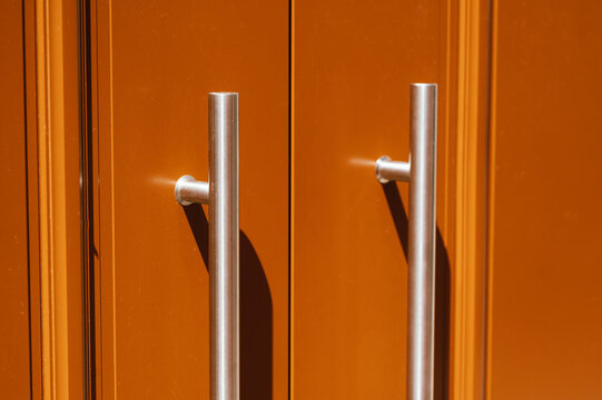 Close-up view of two vertical stainless steel tubular door pulls mounted on an orange painted double door. The symmetrical handles cast soft shadows on the