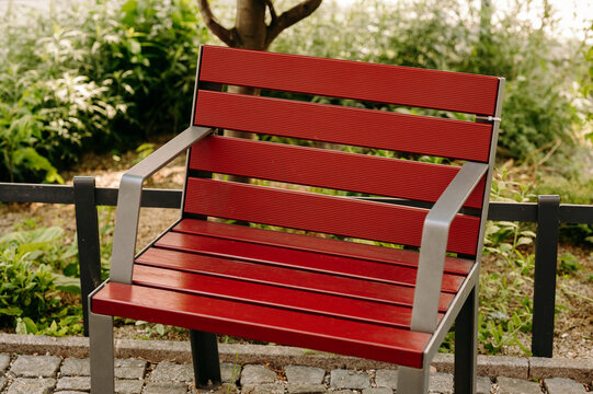 Close-up view of a red slatted wooden bench with gray metal armrests and legs positioned on a cobblestone path beside a low metal railing. Soft natural light