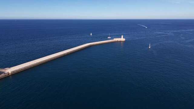 Aerial view of Saint Elmo lighthouse at the end of a long stone breakwater with sailboats on the deep blue Mediterranean Sea in Valletta, Port Region, Malta.