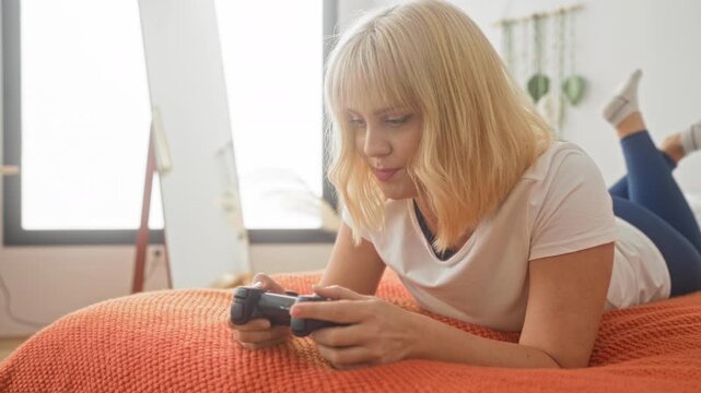Blonde woman holding game controller with bare hands lying on orange bedspread on bed in bedroom with mirror nearby; relaxed leisure concentration.