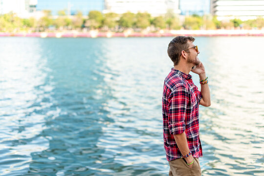 Young man in plaid shirt standing by the waterfront, looking thoughtful.