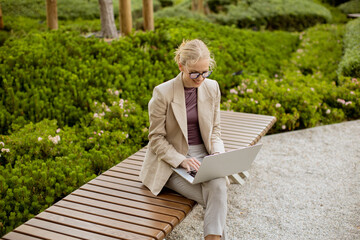 Woman works on laptop in park surrounded by green plants and flowers in daytime © BGStock72